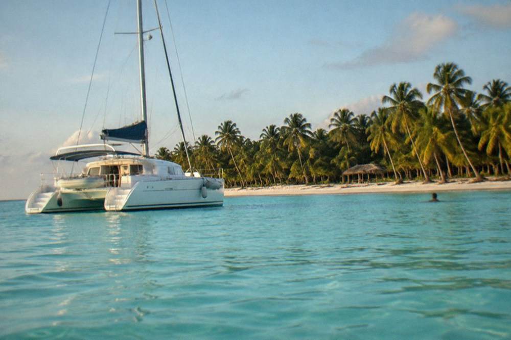 A catamaran anchored in shallow turquoise water off a palm-lined white sand island.