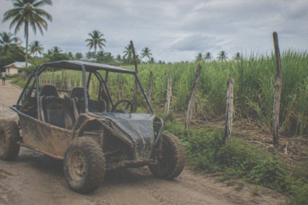 An open-air buggy parked on a dirt road beside green farmland and a rustic wooden fence.