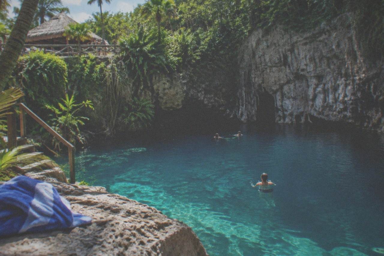 Turquoise freshwater pool at the base of a limestone cliff surrounded by dense tropical vegetation.