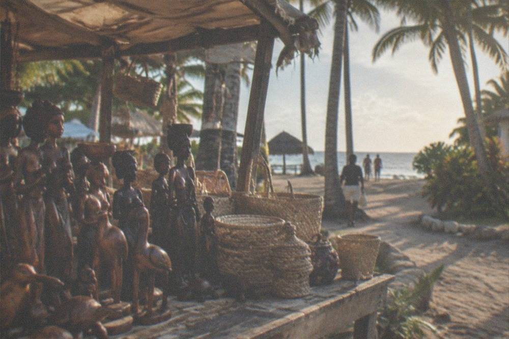 A quiet souvenir market stall shaded by a canopy with carved wooden figures and woven goods on display.