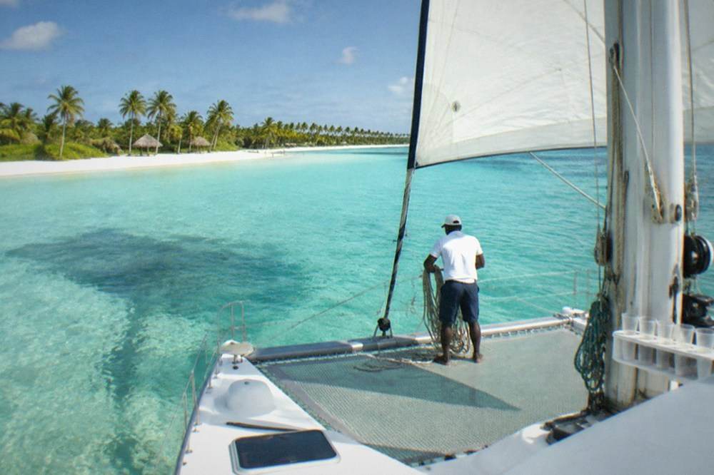 Catamaran with sails up anchored near a sandbar in shallow turquoise water