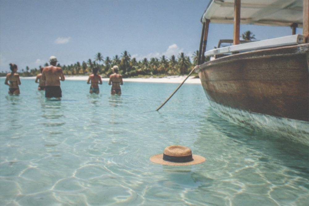 Guests standing in waist-deep clear water on a sandbar with a wooden boat nearby