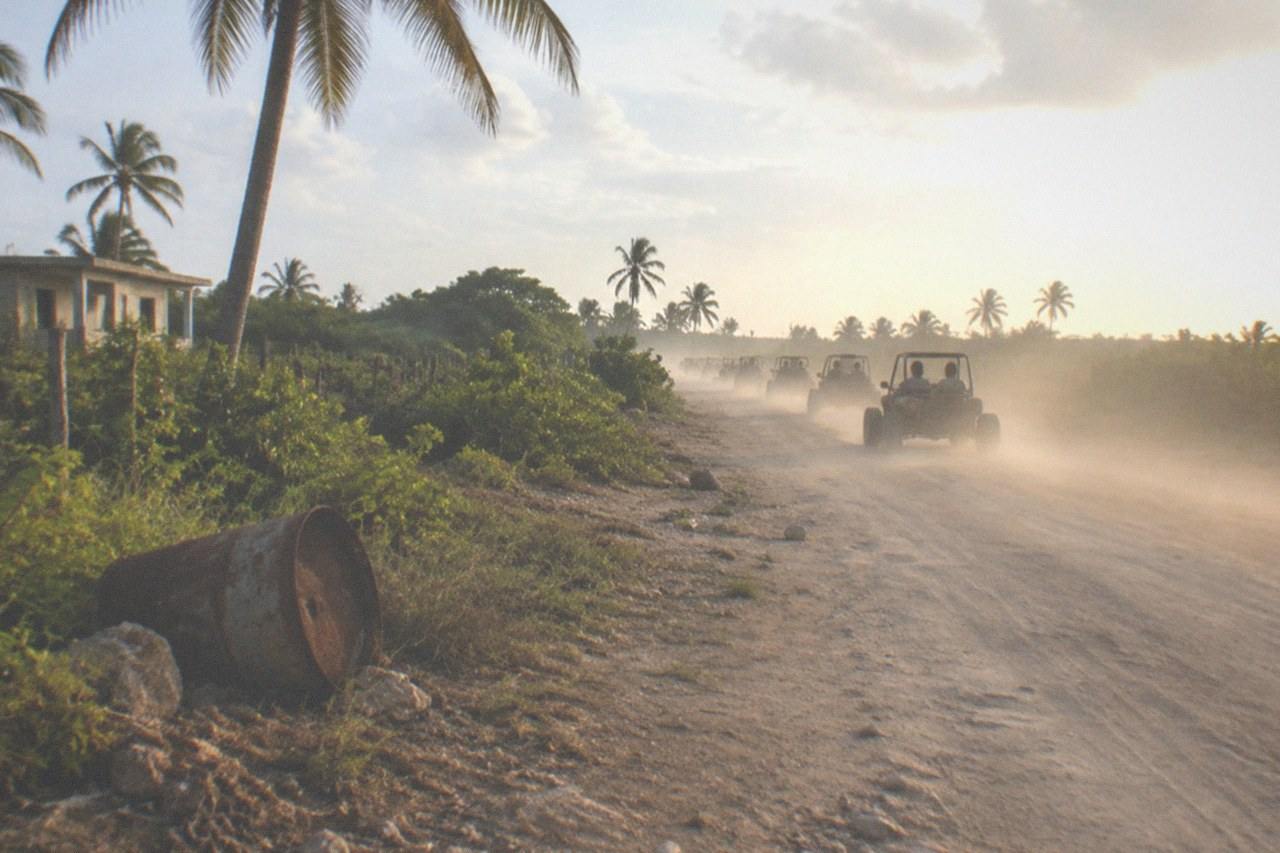 A dirt trail cuts through dense tropical brush, a line of buggies raising a long dust plume as it disappears around a bend ahead.