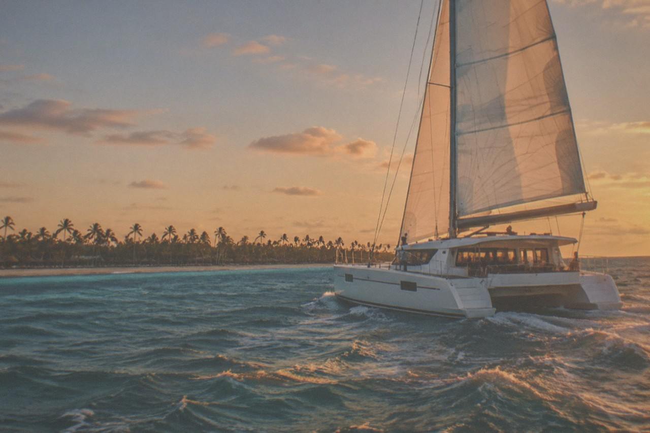 A catamaran under full sail moves along the coastline at golden hour, the low sun casting warm light across the deck and open water.
