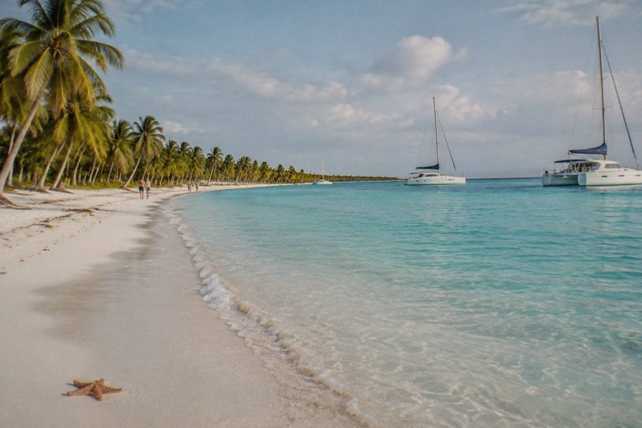 A broad sandbar of white sand fringed with palms stretches into pale turquoise sea, several catamarans anchored in the shallows beyond it.