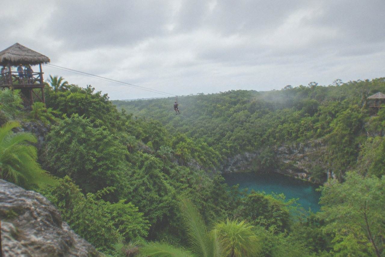 Wide view across a green jungle canopy with zipline cables strung between distant wooden platforms, the blue pool of a cenote below.